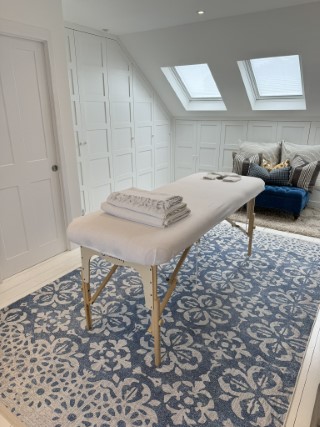 A massage table, with a clean white towelling cover.  The table is on a large blue and white rug. A blue ottoman in the background.